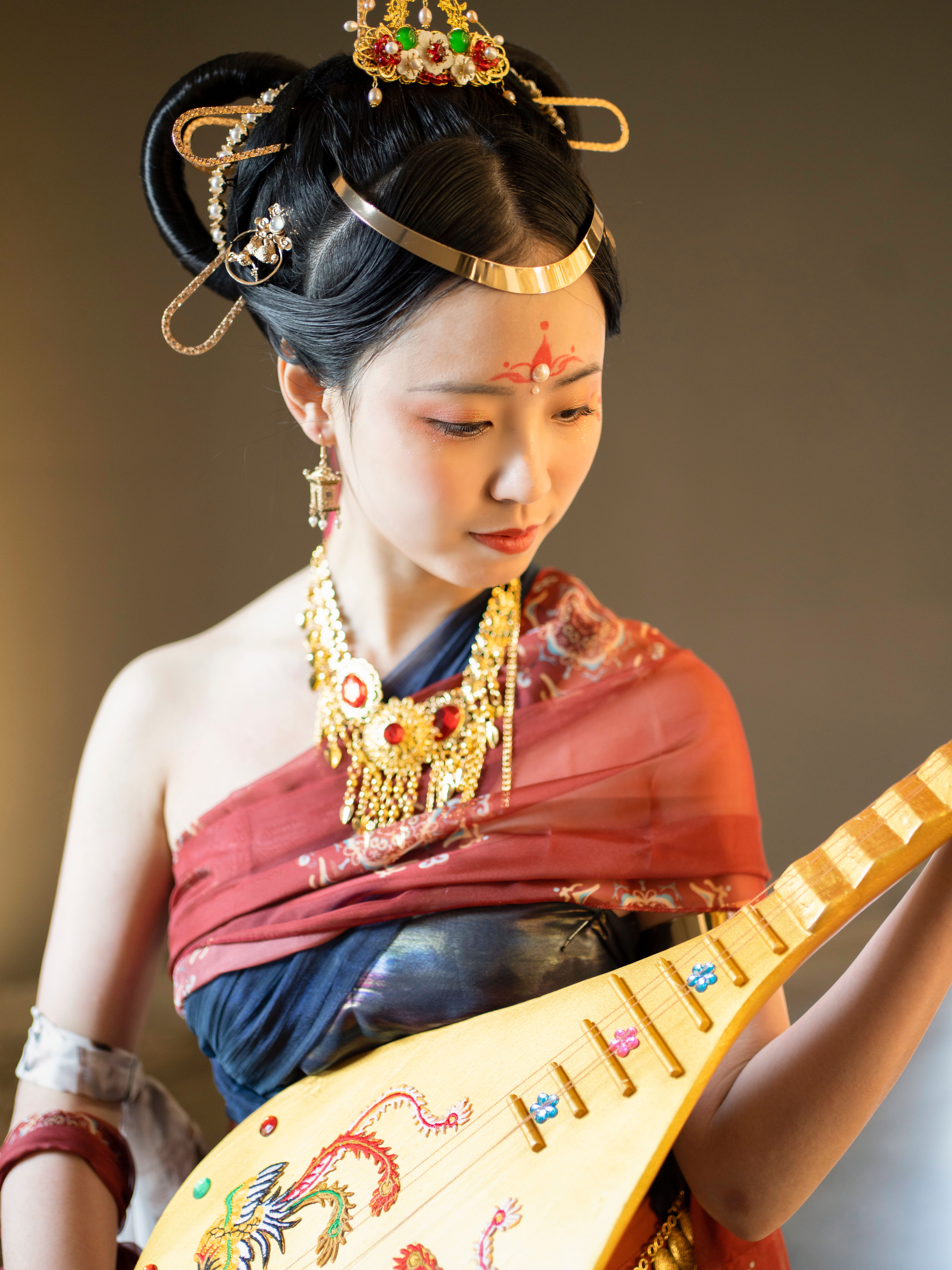 Woman in traditional attire playing a pipa, adorned with gold jewelry, showcasing Asian heritage.