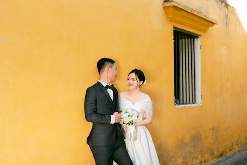 Man in Black Suit Jacket and Woman in White Wedding Dress Standing Beside a Yellow Concrete Wall