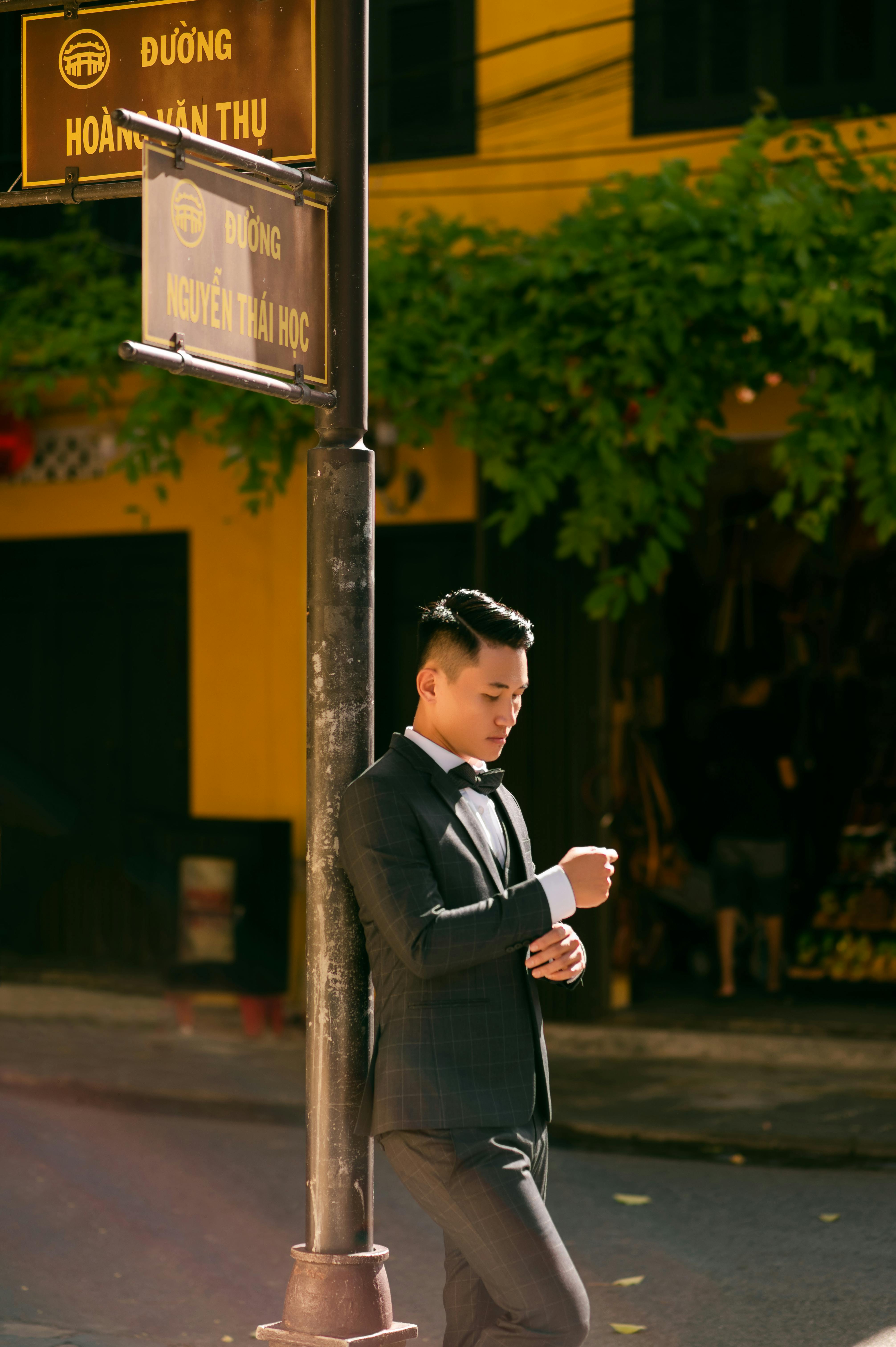 Stylish man in formal attire poses on a sunlit street corner with a road sign, exuding confidence.
