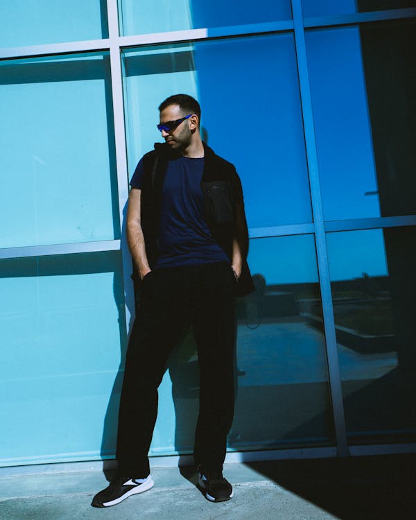 Young Man in a Casual Outfit and Sunglasses Standing Outside of a Modern Building in Sunlight 