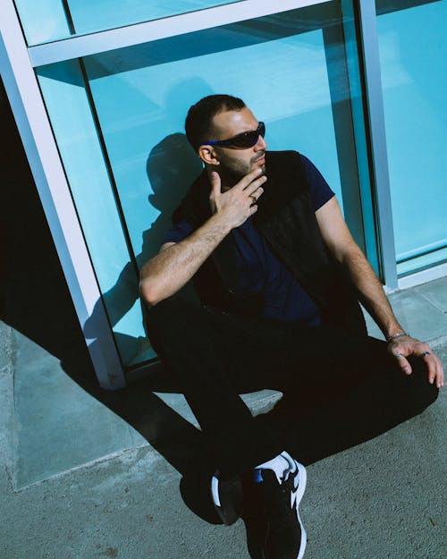 Man in a Casual Outfit and Sunglasses Sitting on the Ground with His Back against a Glass Facade