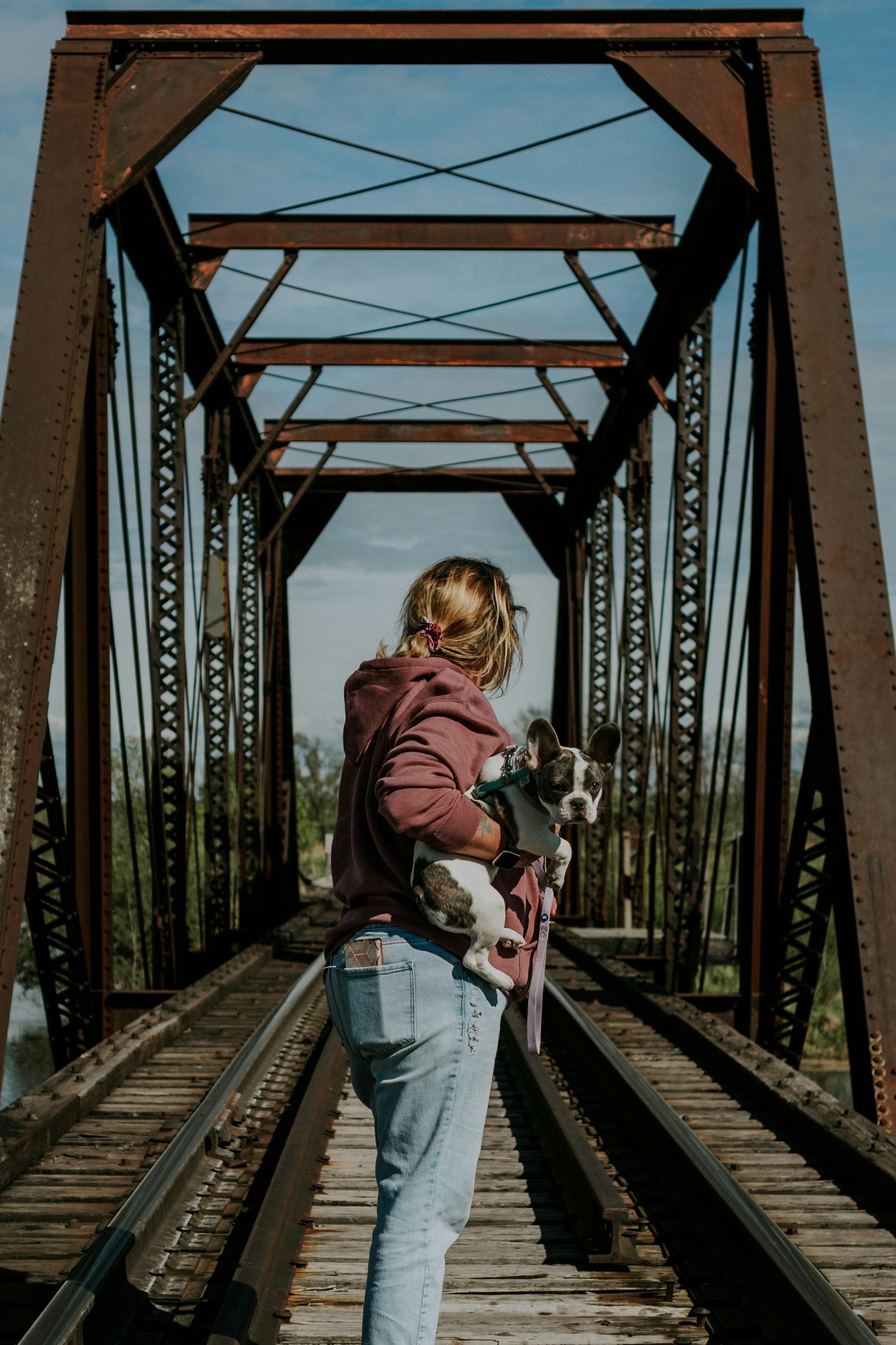 A woman holds her dog as she walks across a vintage railway bridge, showcasing steel structure.