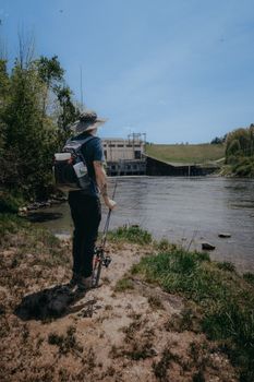 A man standing by a river with fishing gear on a sunny day, enjoying nature.