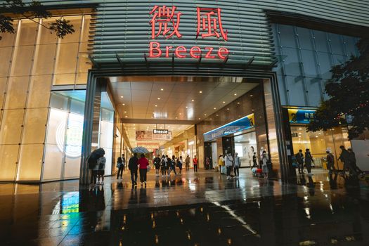 A bustling shopping mall entrance with people and umbrellas on a rainy evening.