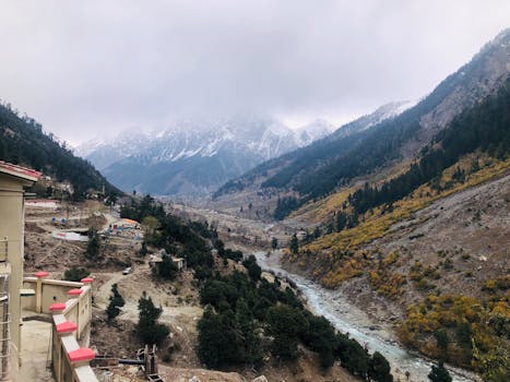 Scenic mountain valley with mist and snow-capped peaks, surrounded by autumn foliage and flowing river.