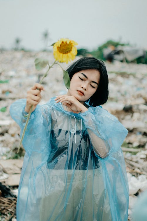 Young Woman Wearing a Raincoat, Holding a Sunflower and Standing on a Field 