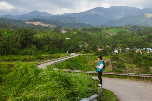 A young man takes a photo of a lush green mountain landscape on a sunny day.