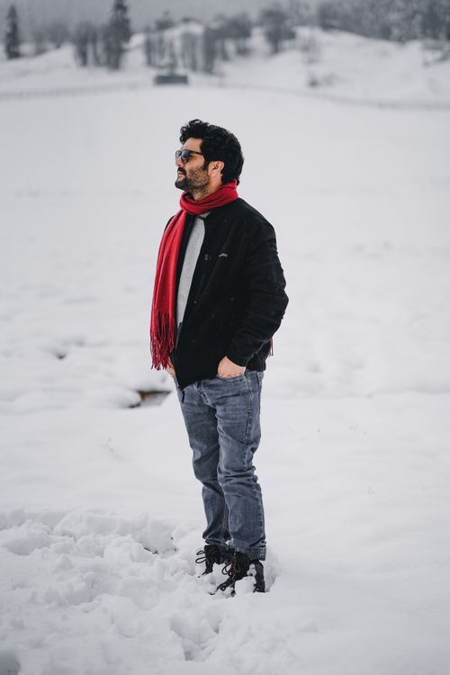 Free A man dressed warmly stands amidst a snow-covered field, wearing a red scarf and black jacket. Stock Photo