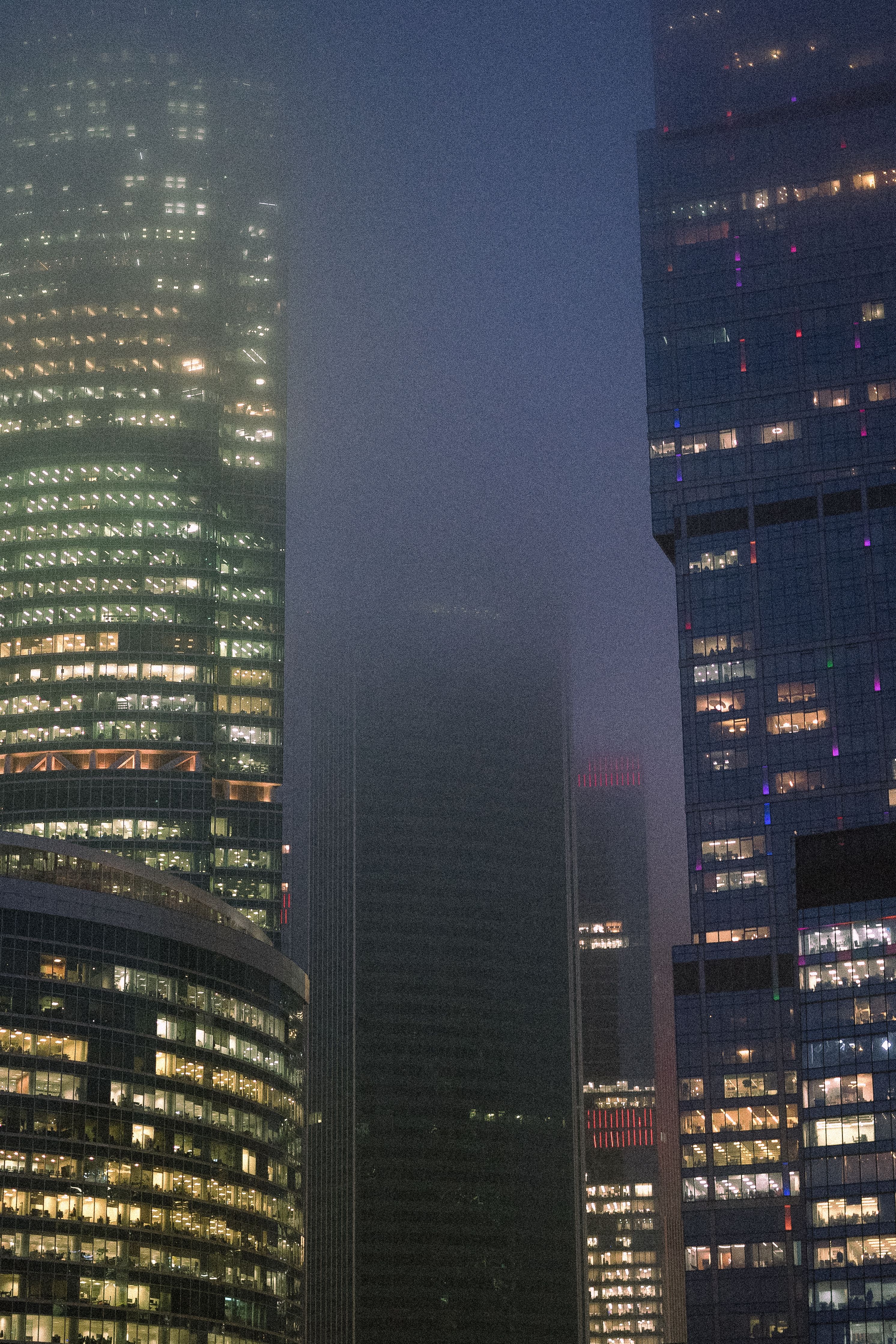 A foggy night view of illuminated skyscrapers in a bustling urban skyline, exuding a modern city atmosphere.