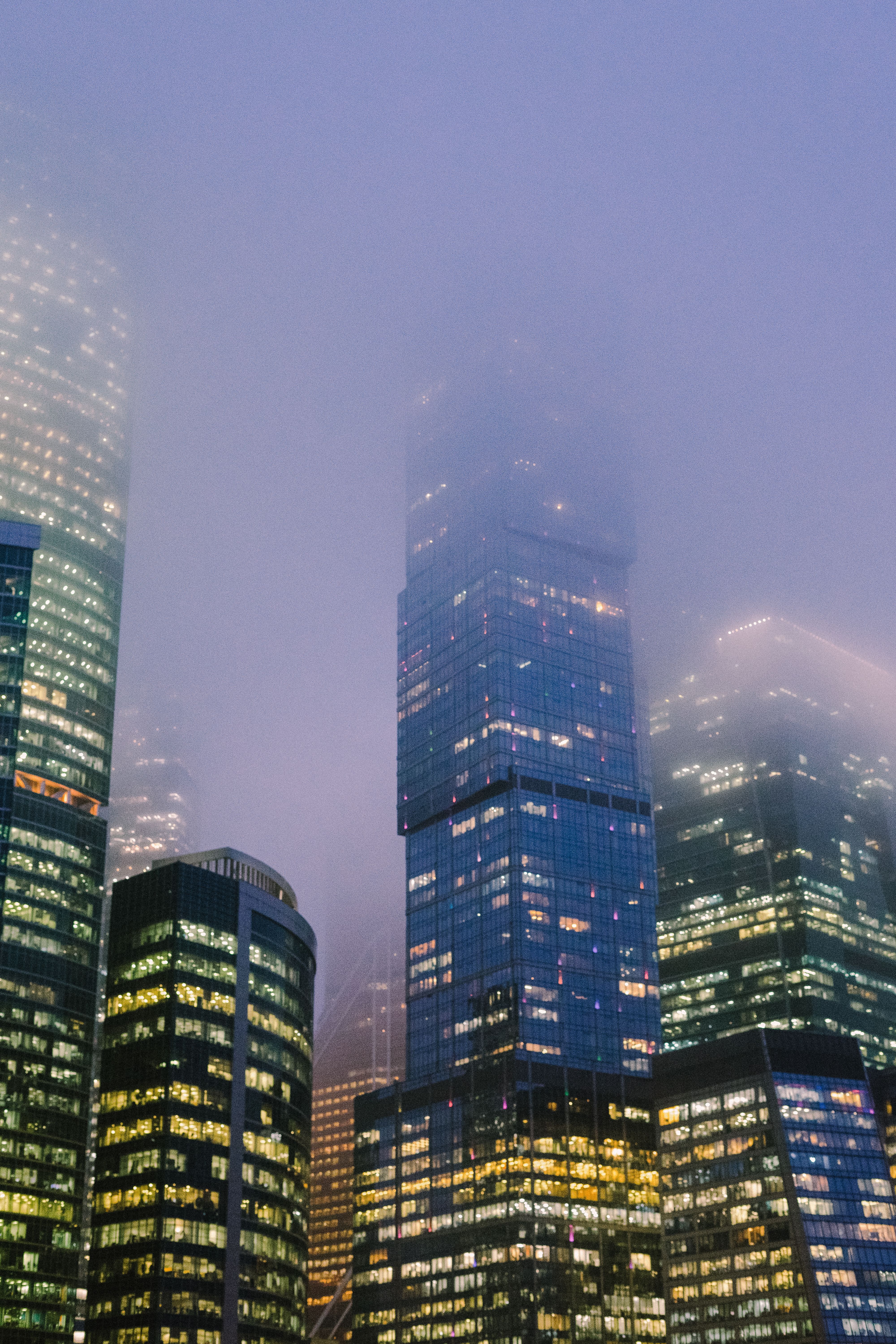 Misty evening view of a modern city skyline with illuminated skyscrapers under foggy conditions.