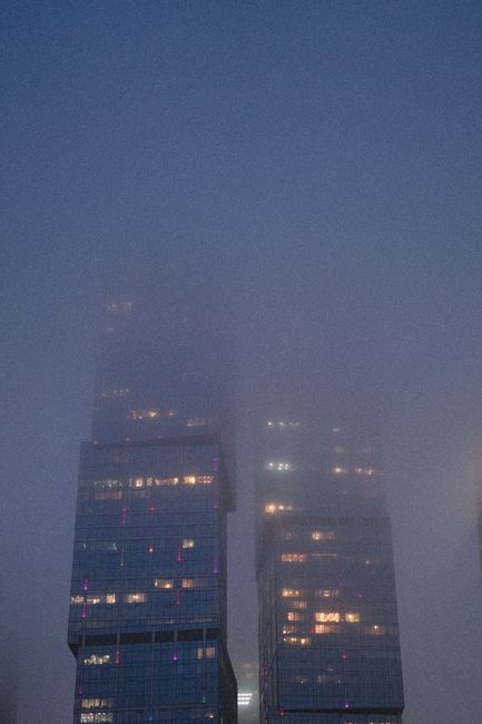 High rise skyscrapers partially hidden by evening fog with illuminated windows, giving a moody urban feel.