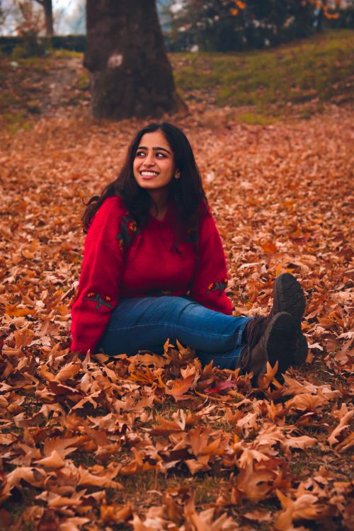 Free Young woman in red sweater sitting in a park, surrounded by autumn leaves, smiling. Stock Photo