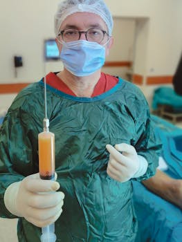 A surgeon in green scrubs, mask, and gloves holding a syringe in a hospital operating room.