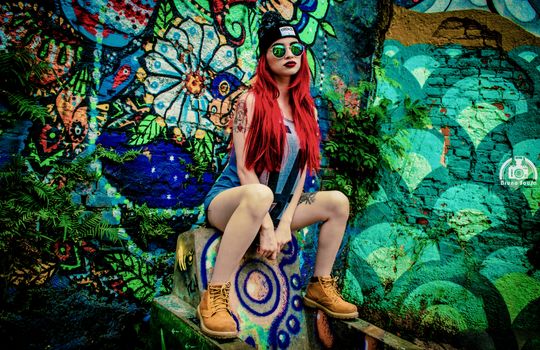 Young woman with red hair and beanie poses fashionably against a colorful graffiti backdrop.