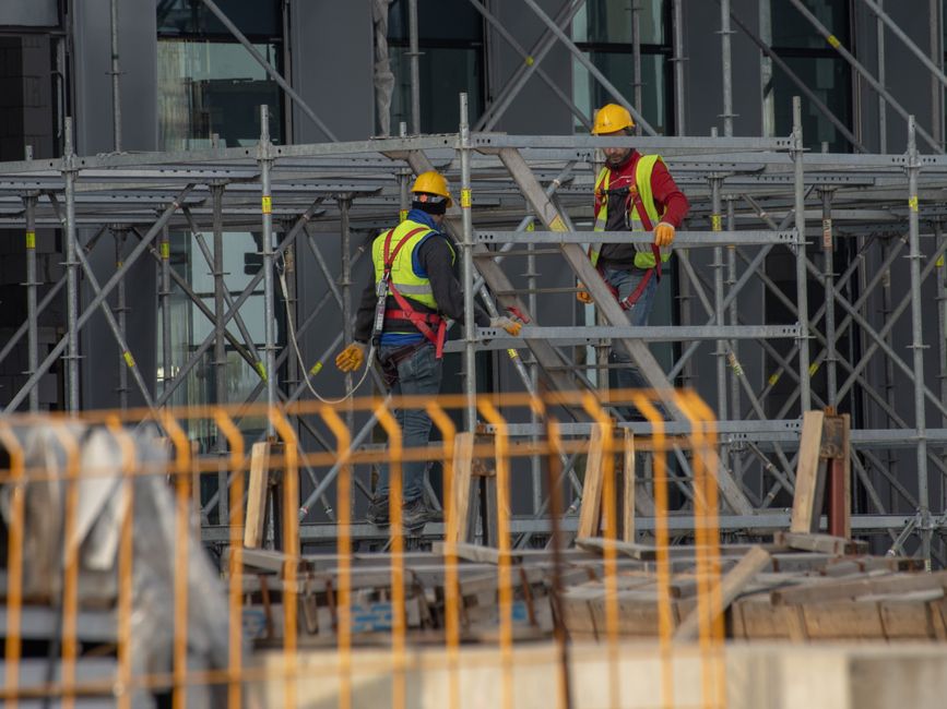 Dos trabajadores de la construcción con equipo de seguridad y cascos trabajando en andamios al aire libre.