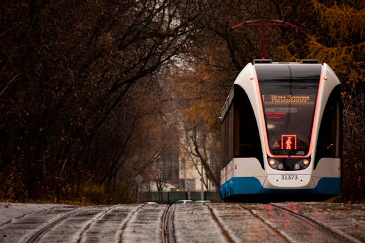A sleek tram travels through a rainy urban street surrounded by autumn trees, evoking a moody atmosphere.