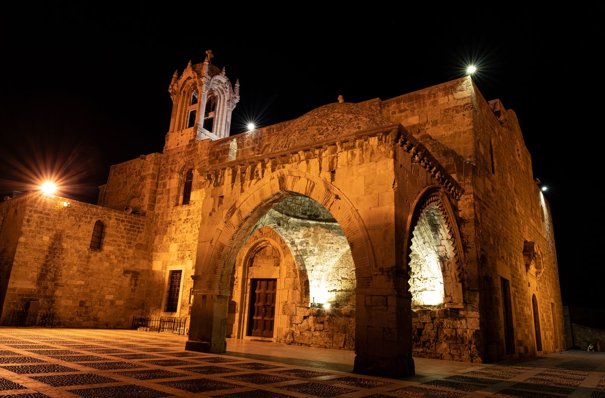The stone facade of the Crusader-era Church of St John the Baptist in Byblos, its layered pointed arches and bell tower lit at night against a dark sky.