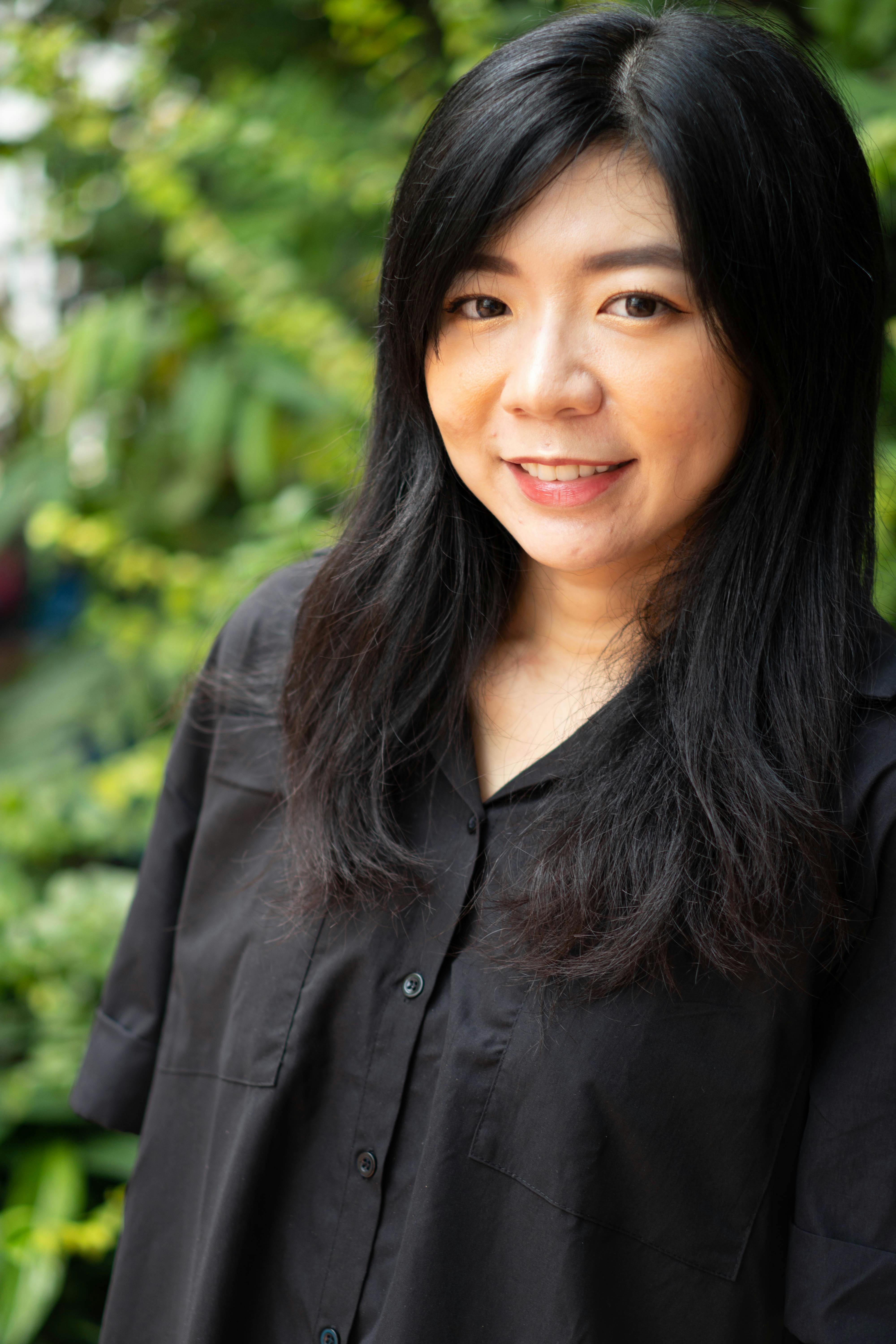 Portrait of a smiling woman with long black hair in a black shirt outdoors against a lush green background.