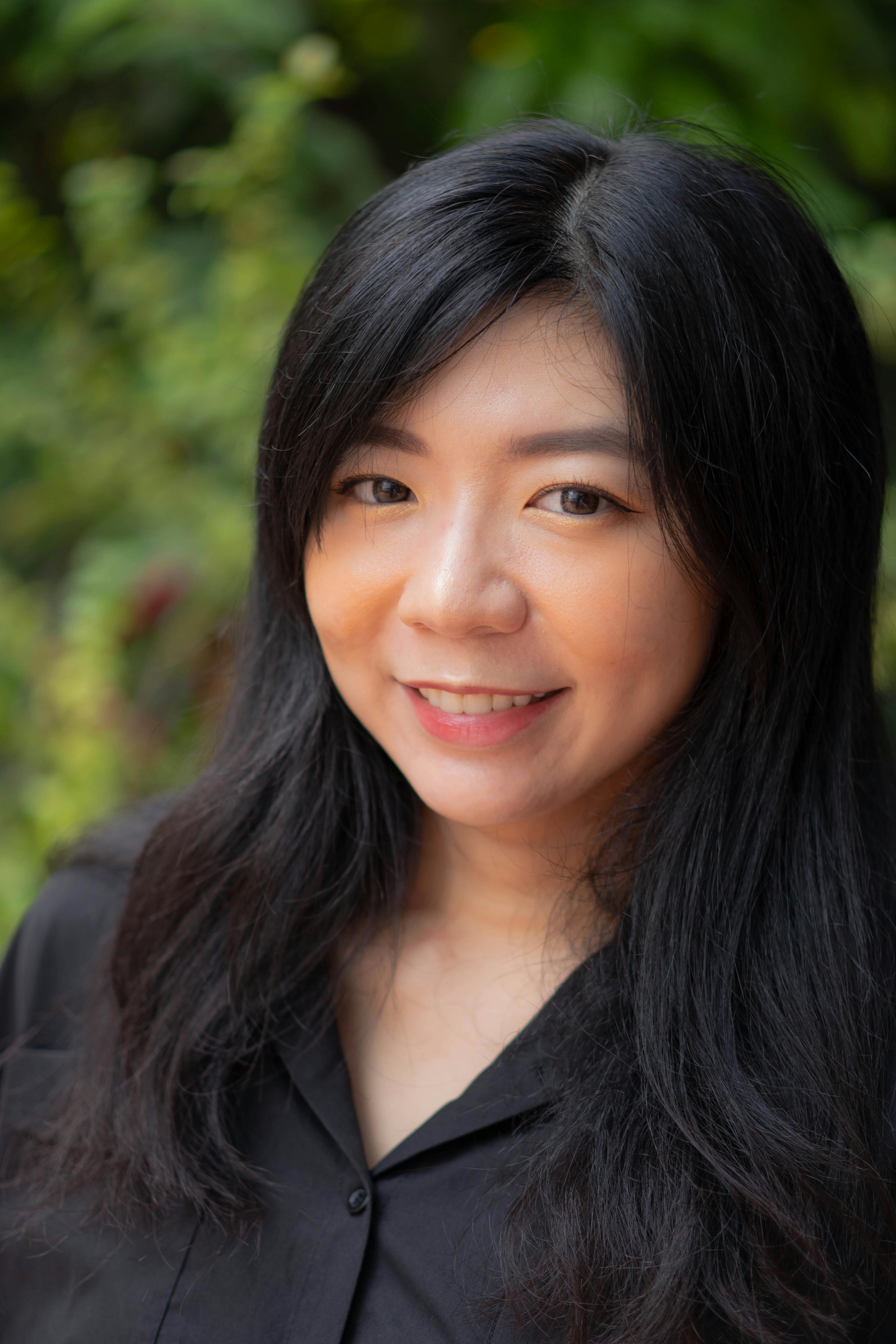 Portrait of an Asian woman with long hair smiling in an outdoor setting, wearing a black shirt.