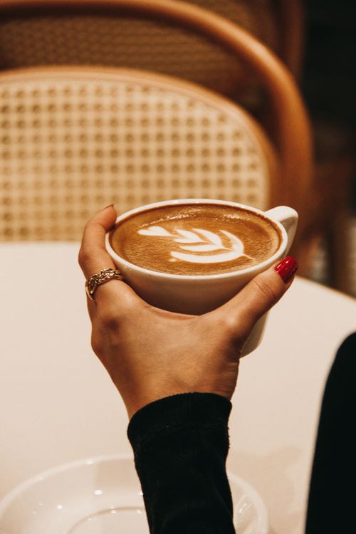 Free A woman's hand holding a cappuccino with delicate latte art in a cozy café ambiance. Stock Photo