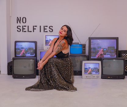 A tattooed woman poses among vintage TVs with a 'No Selfies' sign, embracing retro style.