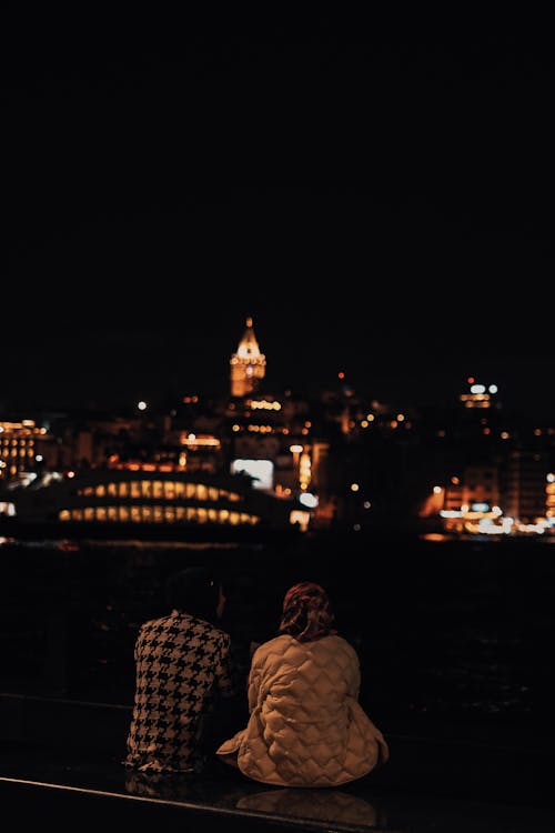 Back View of Two People Sitting on the Shore of the Bosphorus Strait at Night Looking at the Illuminated Buildings of Istanbul 