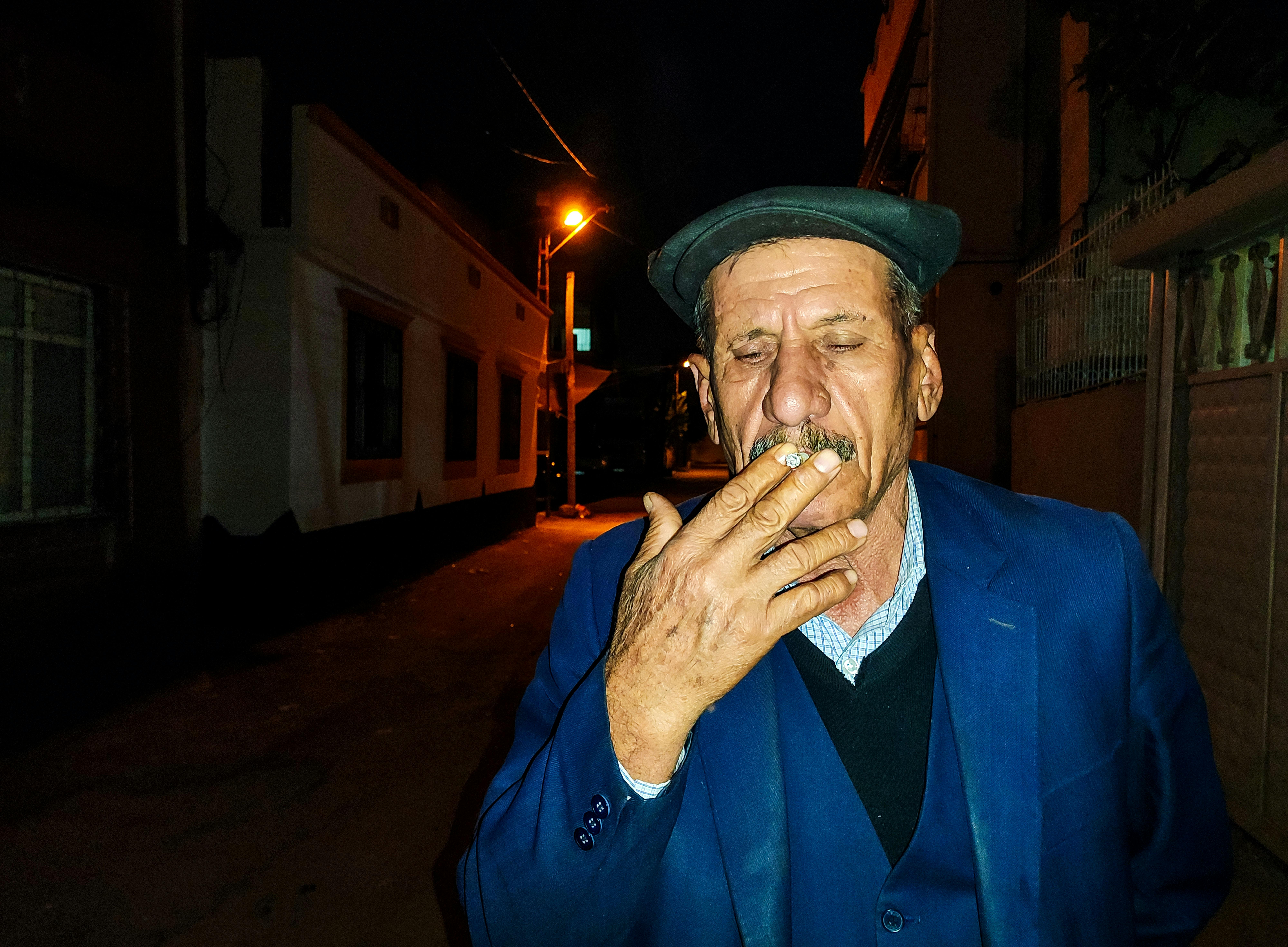 An elderly man in a blue suit smokes thoughtfully on a dimly lit street at night in Adana, Turkey.