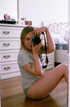 Blonde woman seated on wooden floor, capturing her reflection in a mirror with a camera.
