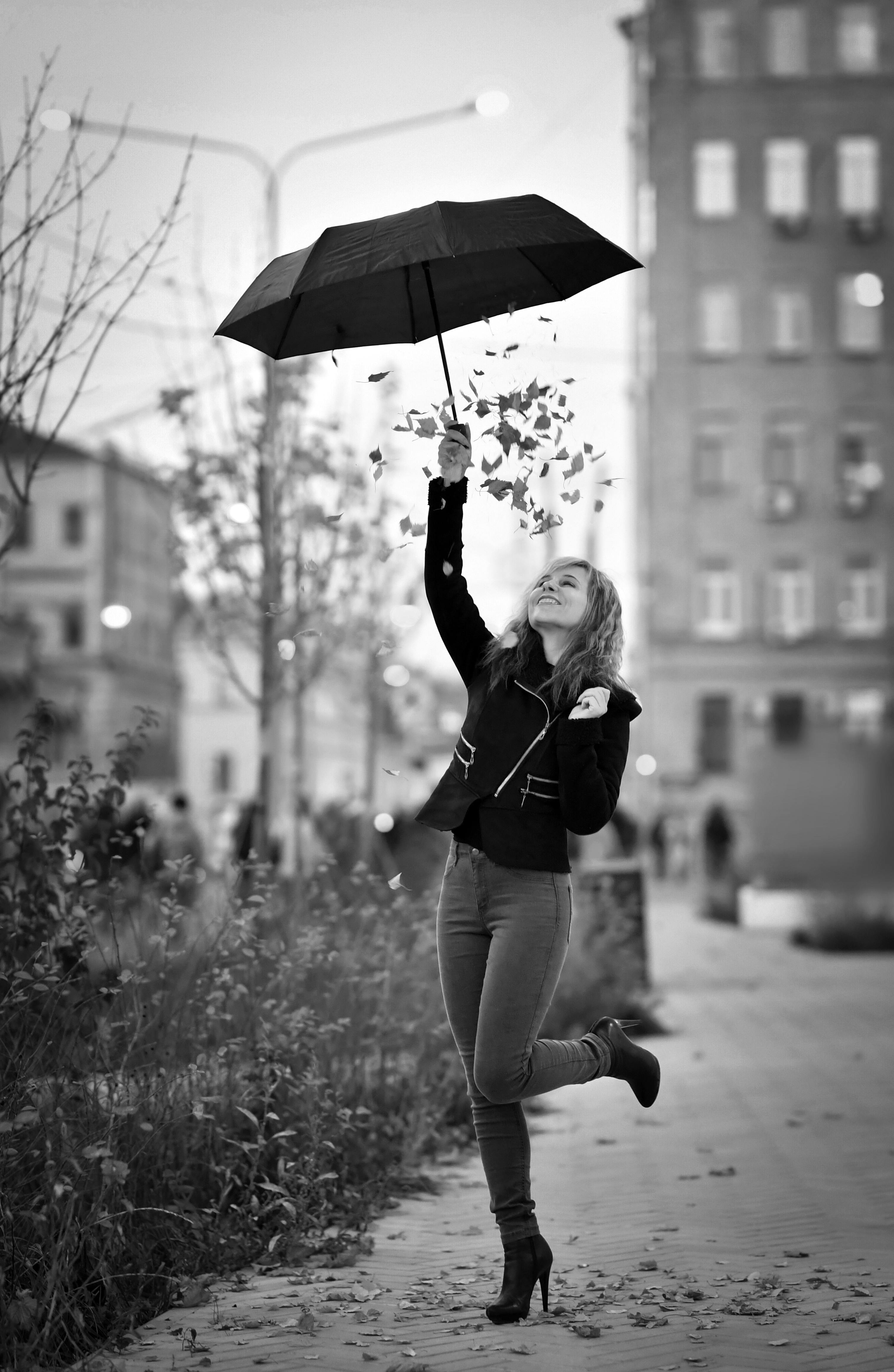 A woman joyfully holding an umbrella amidst falling leaves on a city street.