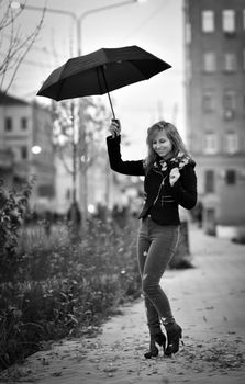 A stylish woman smiles under an umbrella, walking on a rainy street in grayscale.