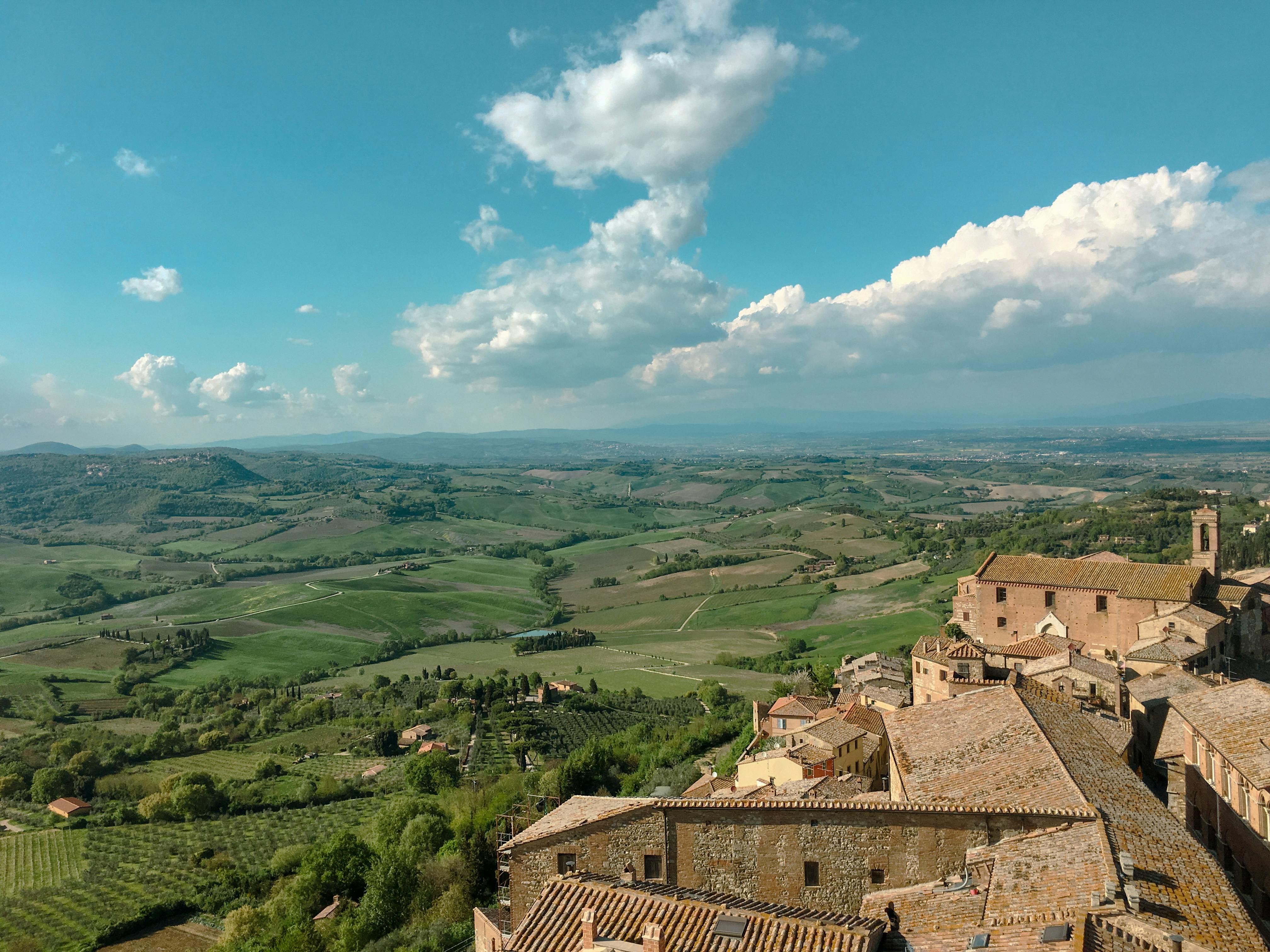 Free Aerial view of Montepulciano, Tuscany, featuring rolling hills, historic buildings, and lush greenery under a clear summer sky. Stock Photo