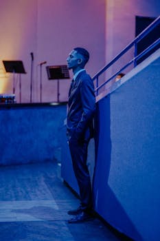 A man in a suit stands indoors near a stage, creating a moody atmosphere.