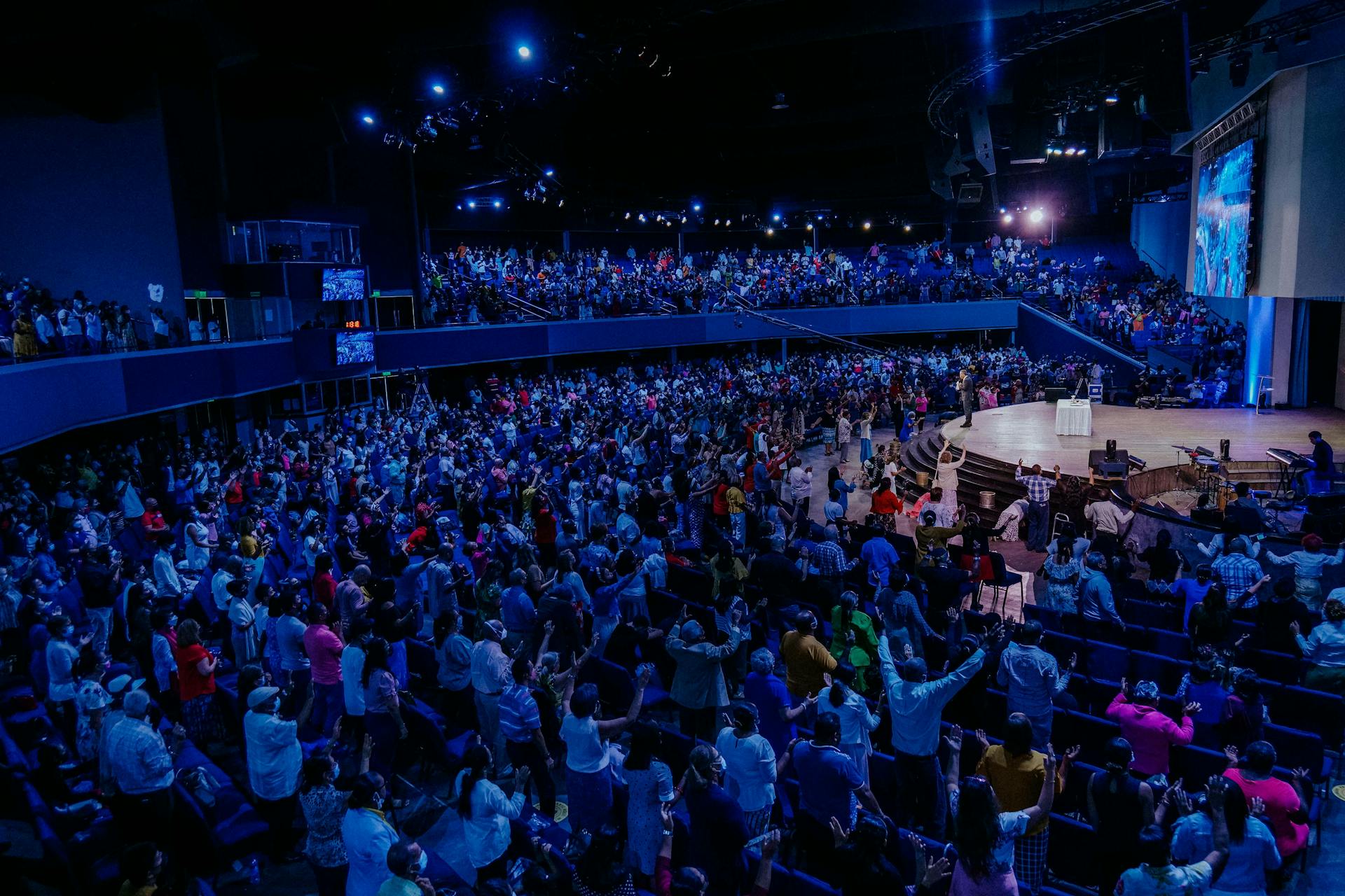 A large vibrant crowd enjoying a live performance in an indoor auditorium under blue lighting.