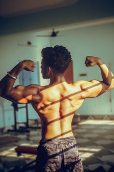 Shirtless man showcasing his muscles while flexing in a gym with natural light.
