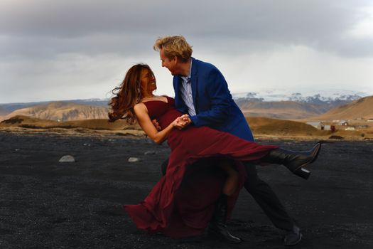 Man in Blue Dress Shirt and Woman in Red Dress Sitting on Gray Sand