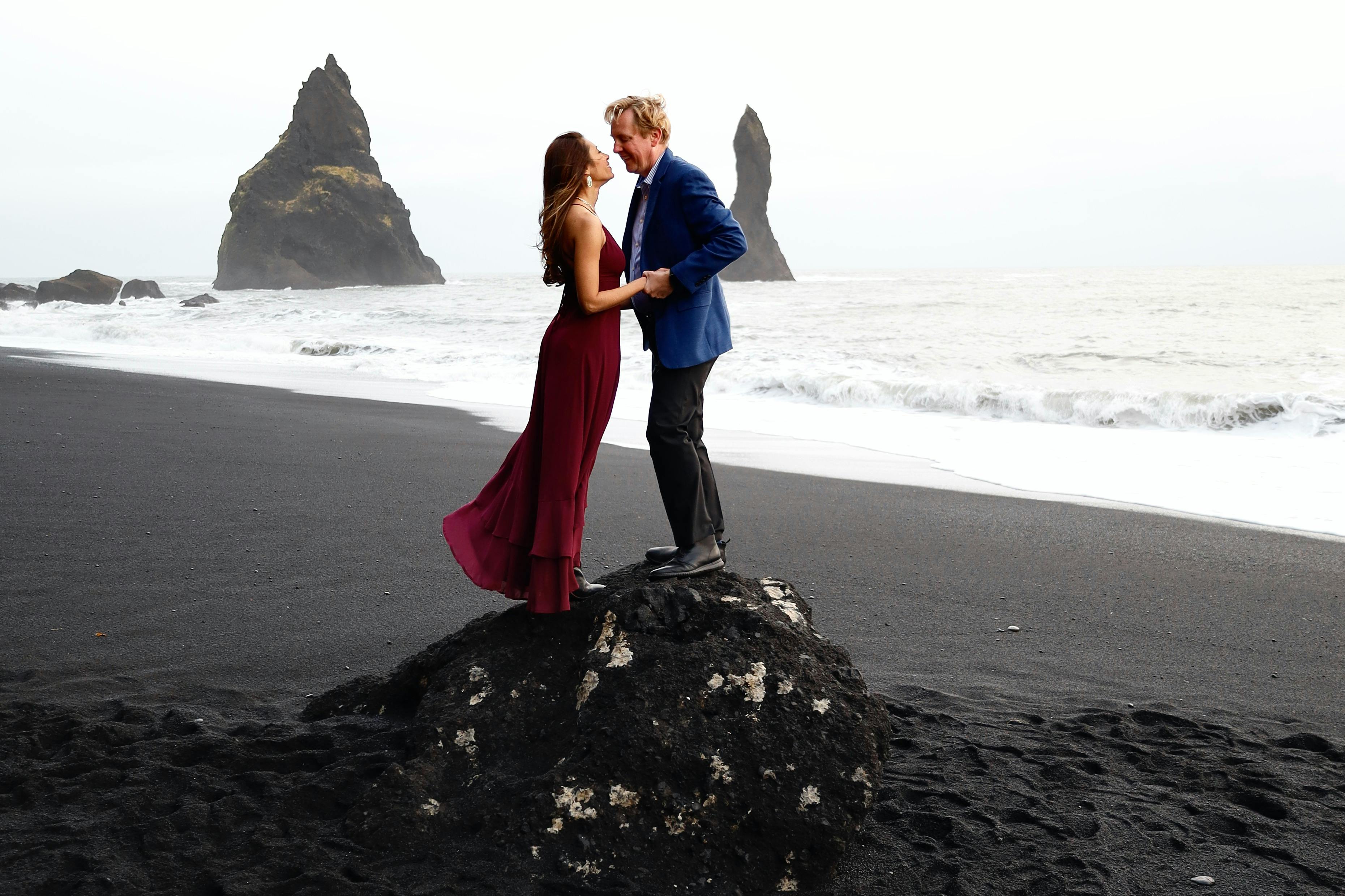 A couple embraces on Reynisfjara Beach, Iceland with dramatic sea stacks in the background. - Vik