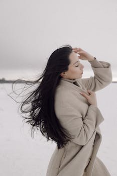 Woman with long dark hair in a beige coat enjoying the wind outdoors against a white sky.