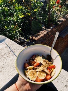 Bowl of cereals with bananas and strawberries held outdoors in sunny weather.