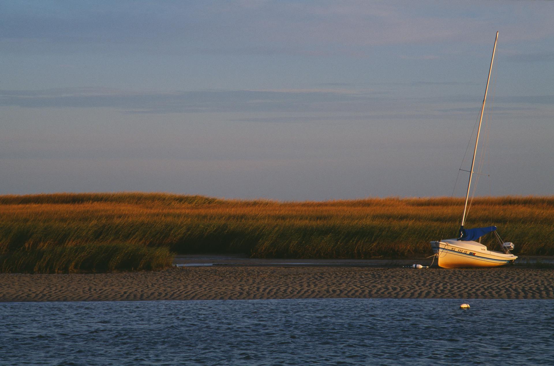 A peaceful scene of a sailboat anchored by the shore at sunset, surrounded by grass fields.