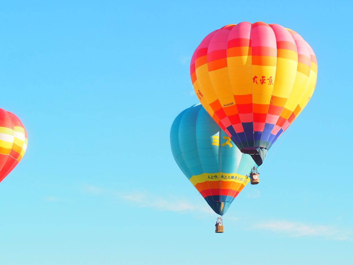 Multiple colourful hot air balloons floating in a clear blue sky — the Saga International Balloon Fiesta competitive flights at dawn