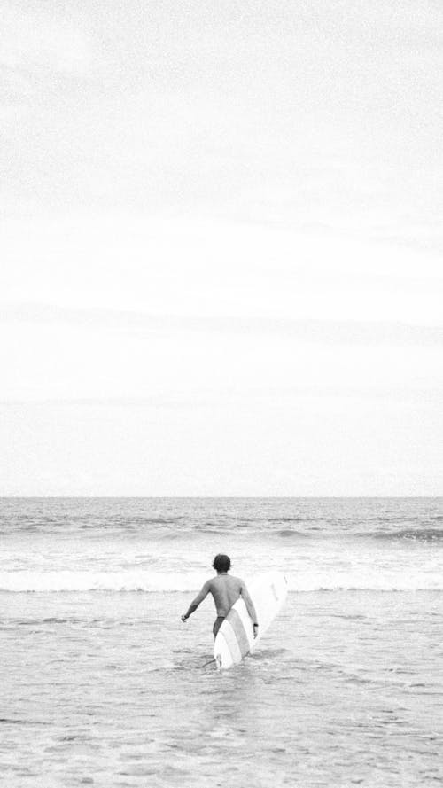 A Grayscale Photo of a Shirtless Man Walking on the Beach while Carrying His Surfboard