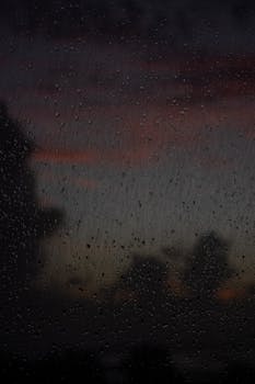 A close-up view of raindrops on a window with a dramatic sunset in the background.
