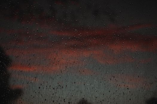 Raindrops on a window with a dramatic sunset background create a moody and introspective scene.