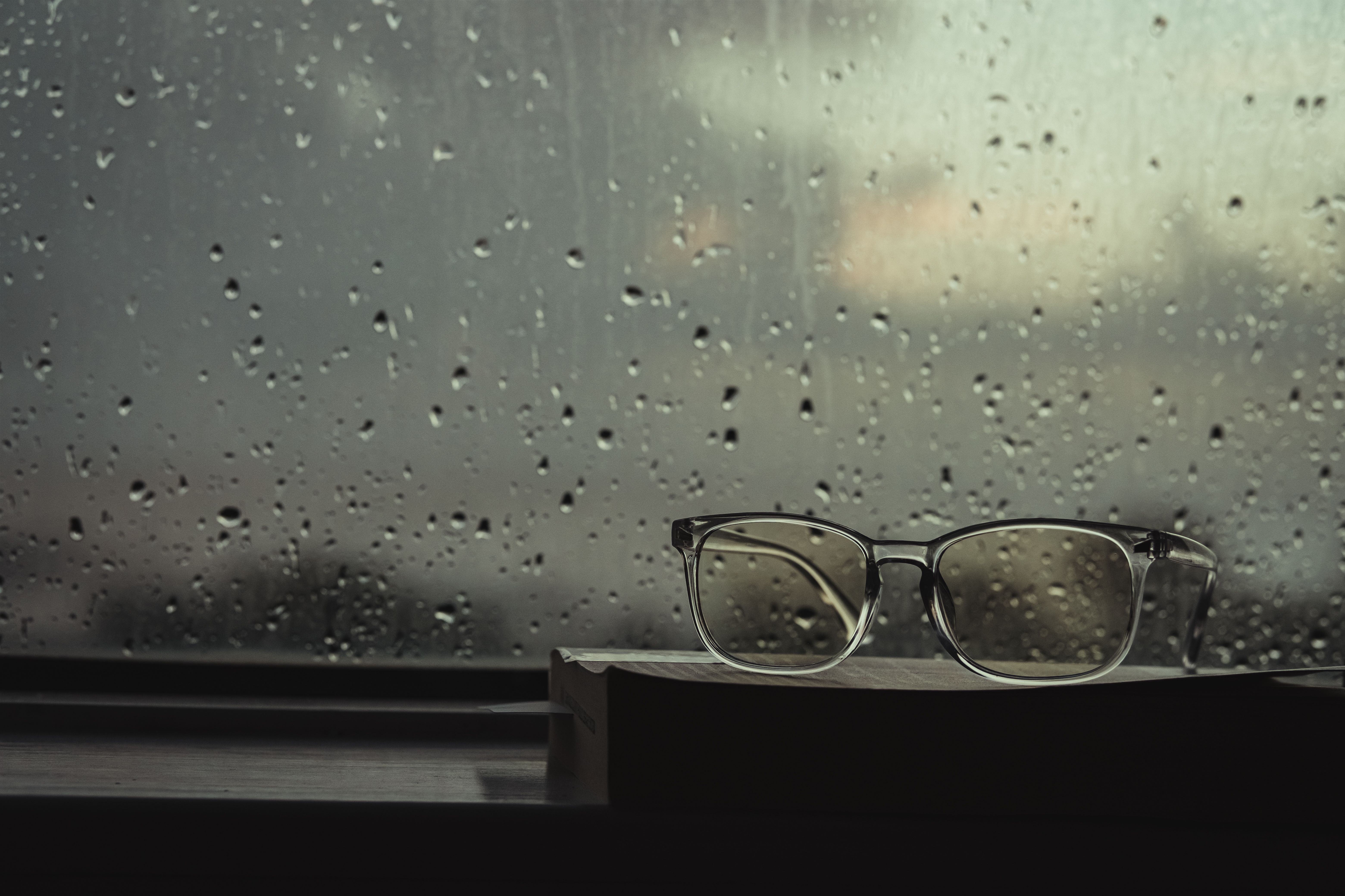 Eyeglasses resting on a book by a window with raindrops. Cozy and reflective mood.