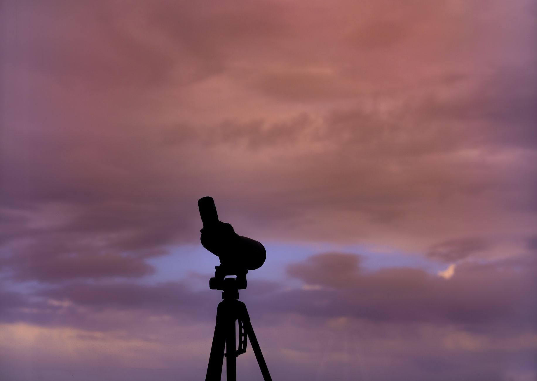 Silhouette of a telescope on a tripod set against a vibrant sunset sky with clouds.