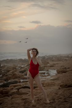 A woman stands on a rocky beach in a red swimsuit during sunset, with birds flying in the background.