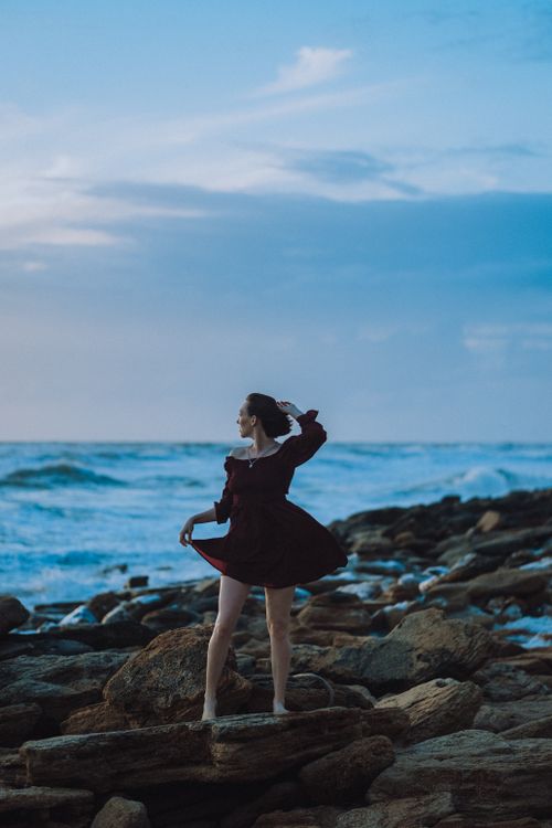 Free A woman stands elegantly on a rocky seashore, gazing at the ocean under a blue sky. Stock Photo