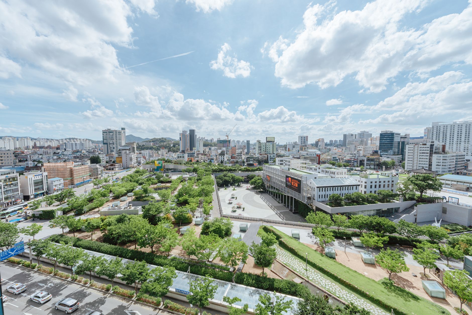 A vibrant cityscape featuring a lush urban park surrounded by modern buildings under a partly cloudy sky.