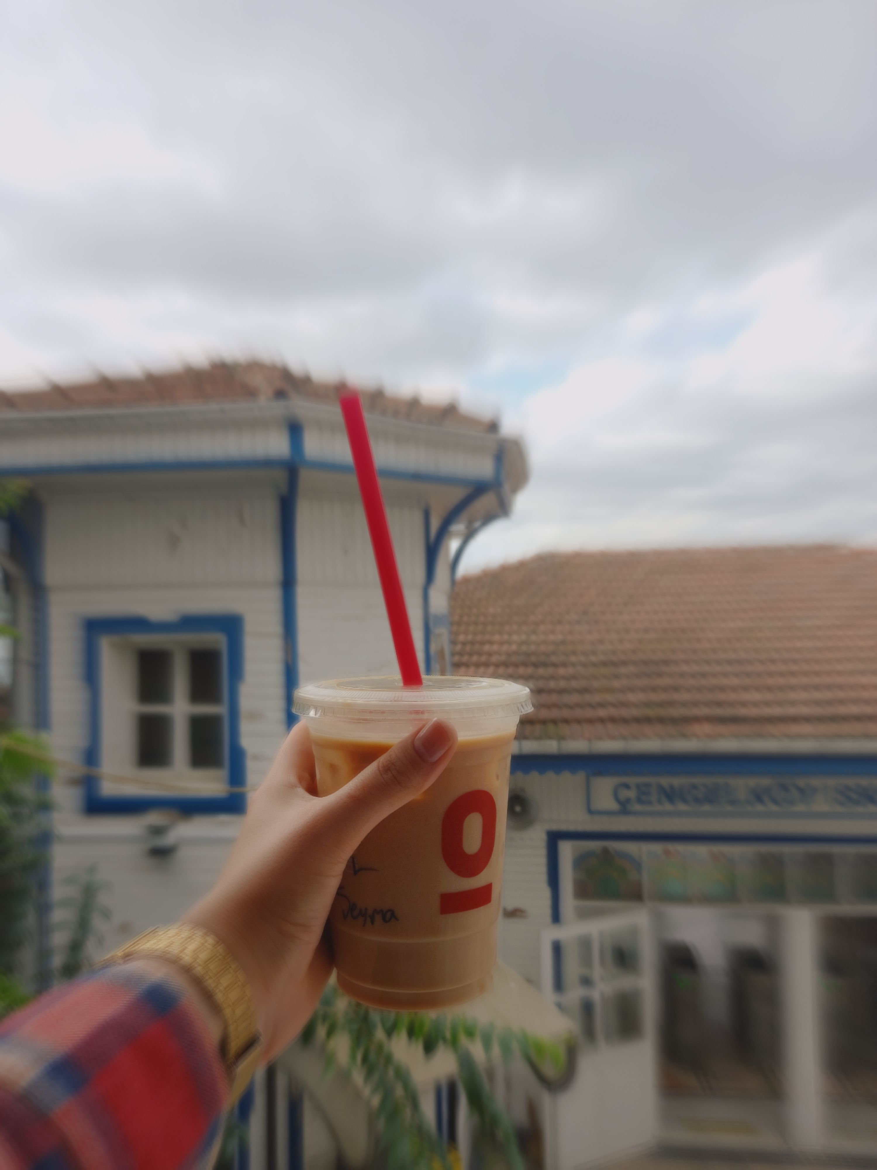 Hand holding iced coffee with red straw, blurred background of vintage building, outdoor scene.