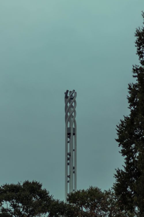 Free A modern sculpture tower rising into a cloudy blue sky with surrounding trees. Stock Photo